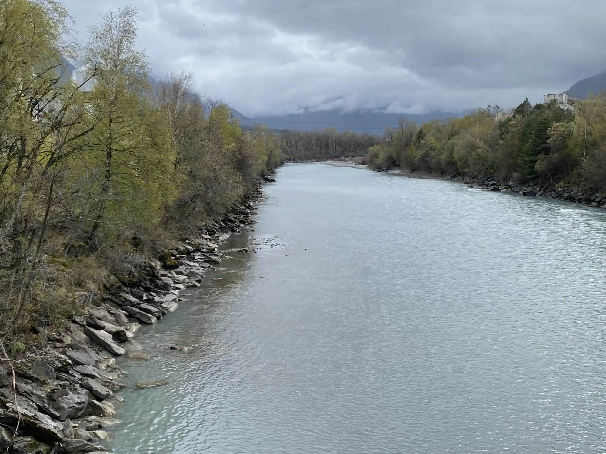 Ein ruhiger Fluss fließt zwischen felsigen, von Bäumen gesäumten Ufern unter einem wolkigen, bedeckten Himmel. In der Ferne sind schwach Berge zu erkennen, die teilweise vom Nebel verdeckt sind.