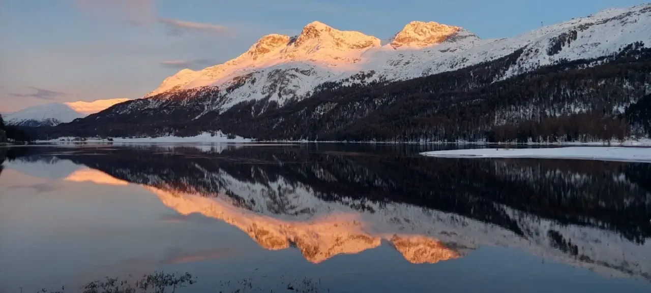 Bei Sonnenaufgang oder Sonnenuntergang sind die schneebedeckten Berge in warmes Sonnenlicht getaucht, das sich deutlich in der stillen, dunklen Oberfläche eines darunterliegenden Sees mit Bäumen und schneebedecktem Ufer spiegelt.