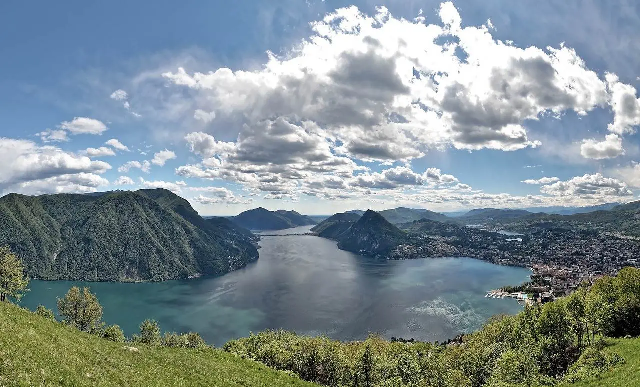 Ein Panoramablick auf den großen Schweizer See, umgeben von grünen Hügeln und einer Stadt. Der Himmel ist teilweise bewölkt, das Sonnenlicht spiegelt sich im Wasser, wo Felchen und Forellen gedeihen und eine malerische und friedliche Landschaft schaffen.